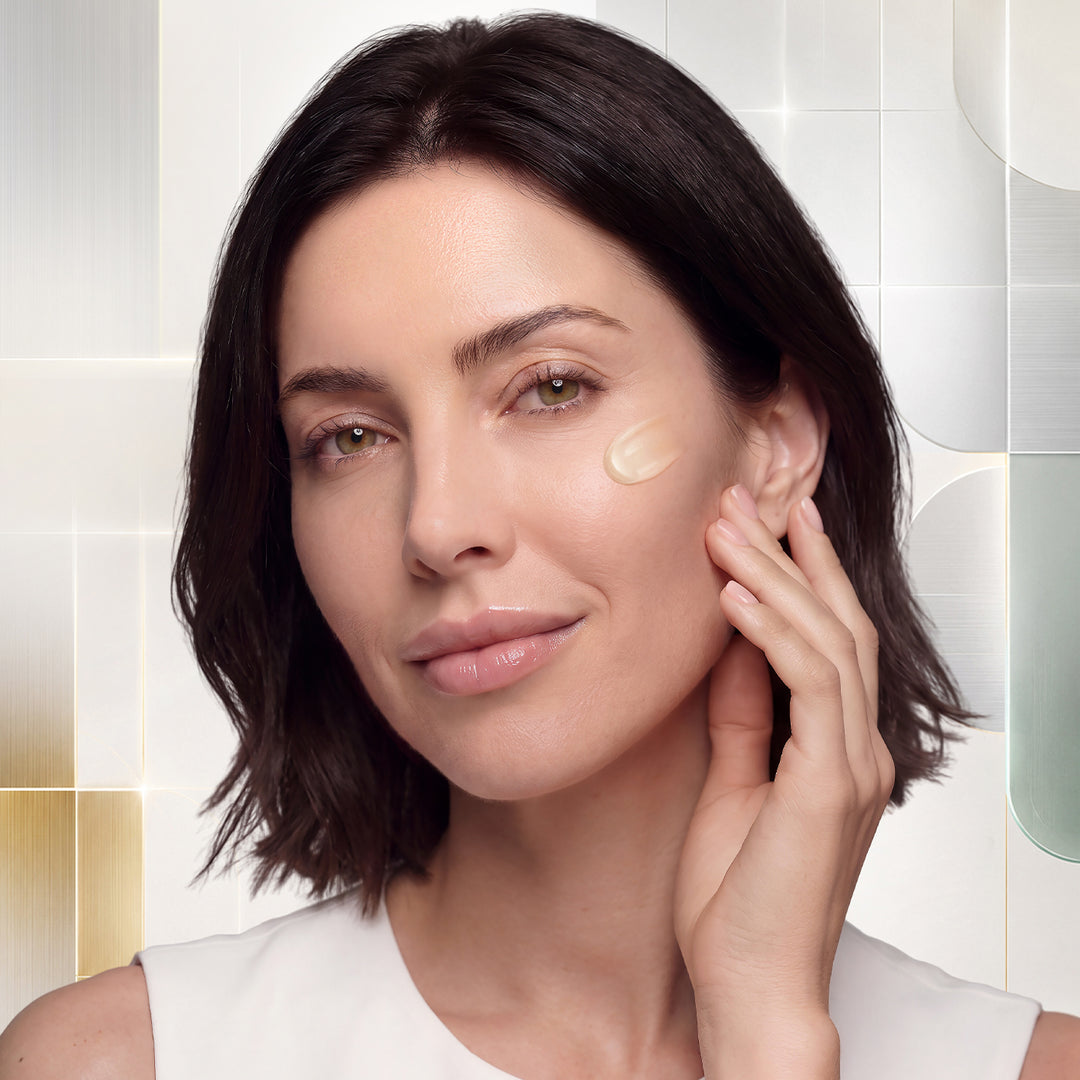 A close-up of a woman with short dark brown hair applying or showing a dollop elixir oil on her cheek. She is wearing a white sleeveless top, set against a modern, softly lit background with geometric shapes.