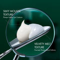 Close-up of a cosmetic tube showing two shades of cream on a silver spatula, under a magnifying lens with text reading 'Silky Mousse Texture' and 'Velvety Melt Texture' on a dark green background.